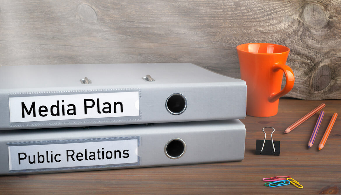 Folders on a desk labeled “media plan” and “public relations”, stacked next to pencils and a coffee mug