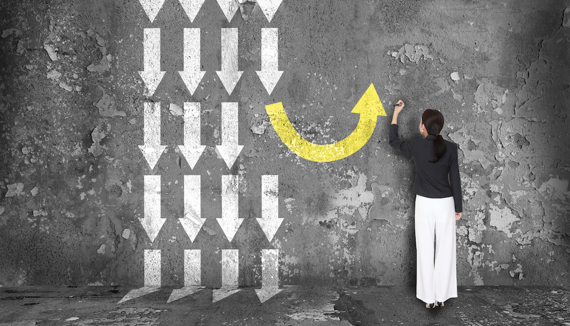 woman in front of black board with lots of white arrows pointing straight down and one yellow arrow curving around to point up, representing making a different decision