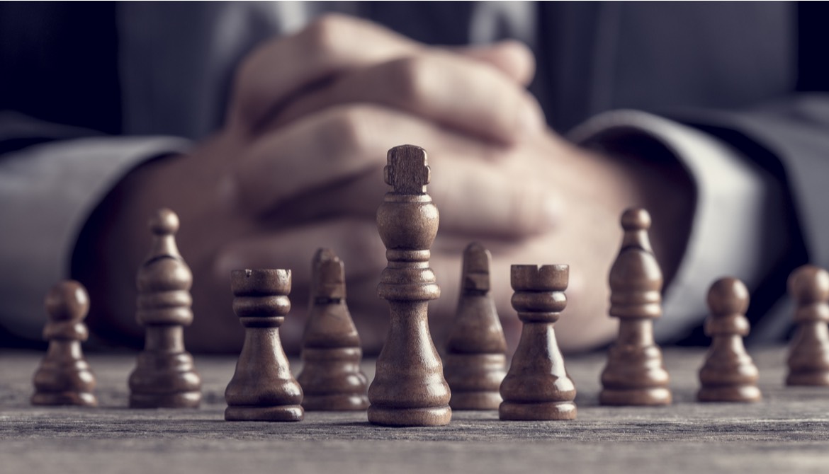 businessman with hands folded behind group of wooden chess pieces