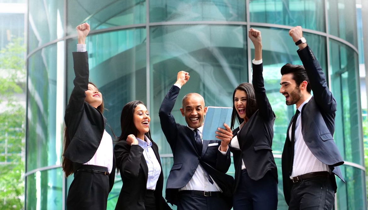 team of cheering colleagues gathered around a coworker holding a digital tablet