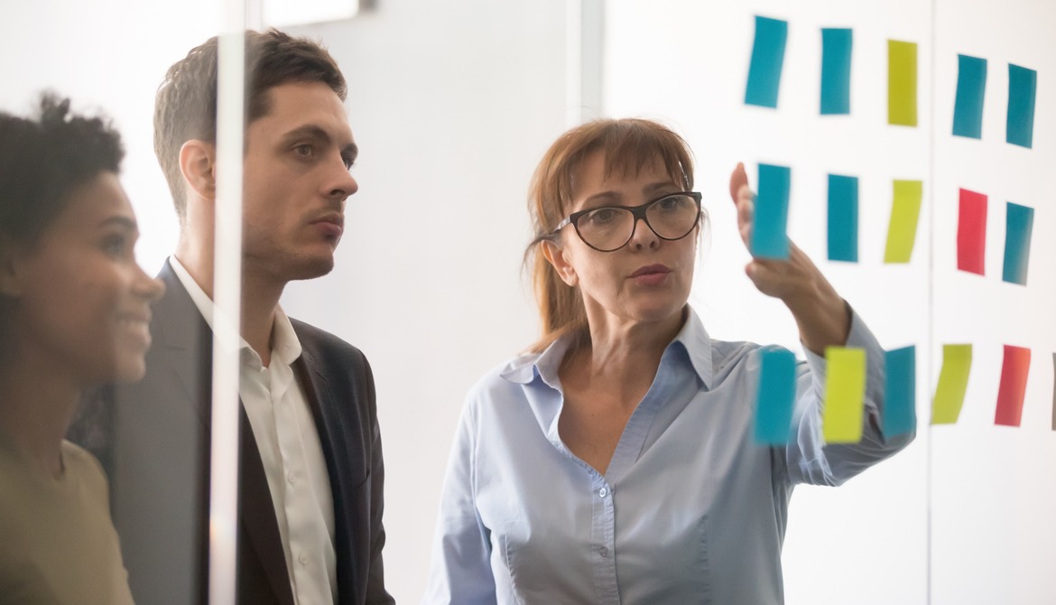 three diverse people discuss information written on colorful sticky notes on a glass wall