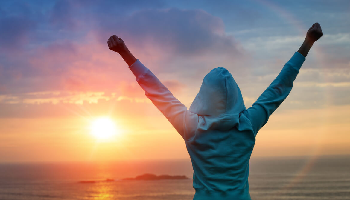 a woman at sunrise at the beach raises arms in victory