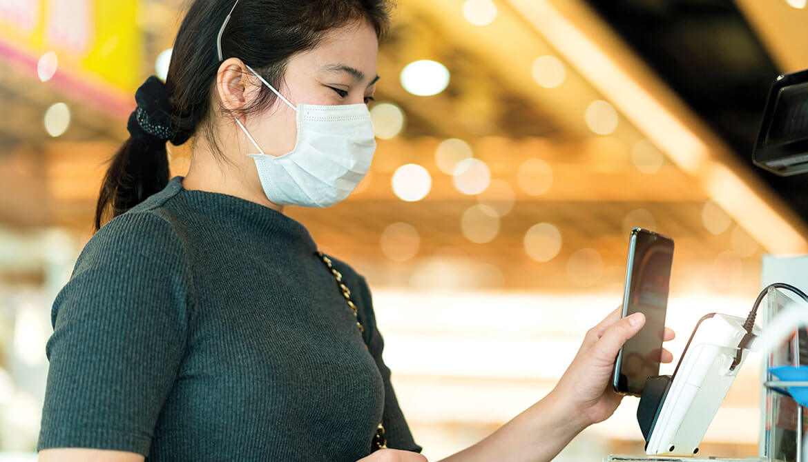 young Asian woman wearing mask holds smartphone up to payment kiosk at store