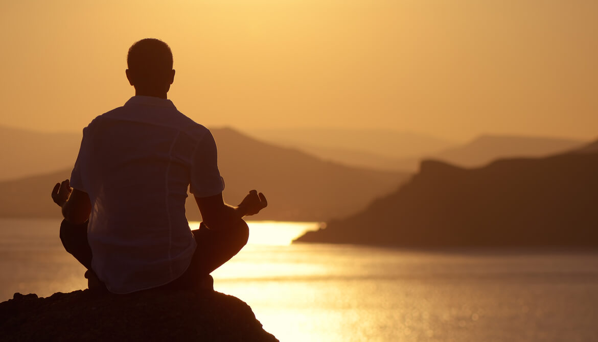 man in white collared shirt sits on rock in yoga position facing water and sunset