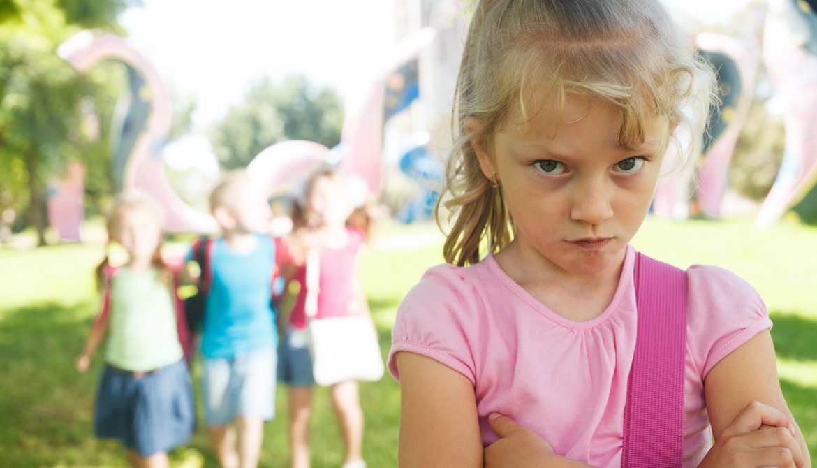 young girl stands by herself with arms folded defensively at playground park while group of 3 children exclude her