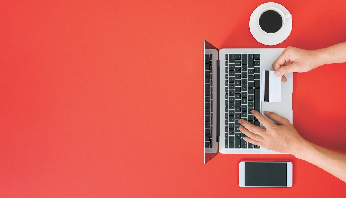 hand holding out credit card while typing information into laptop next to coffee cup and smartphone on red background 