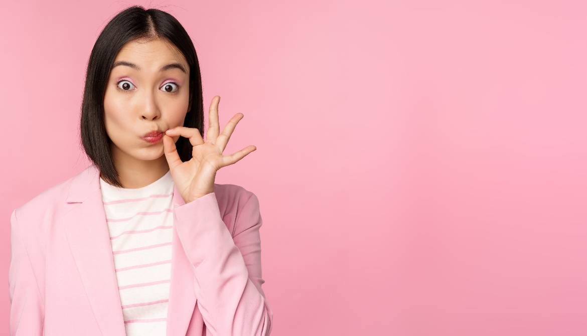 young Asian businesswoman in pink suit pinching lips shut to keep a secret