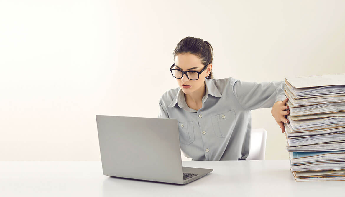 businesswoman working on a laptop while pushing a giant stack of folders and papers off the side of the table