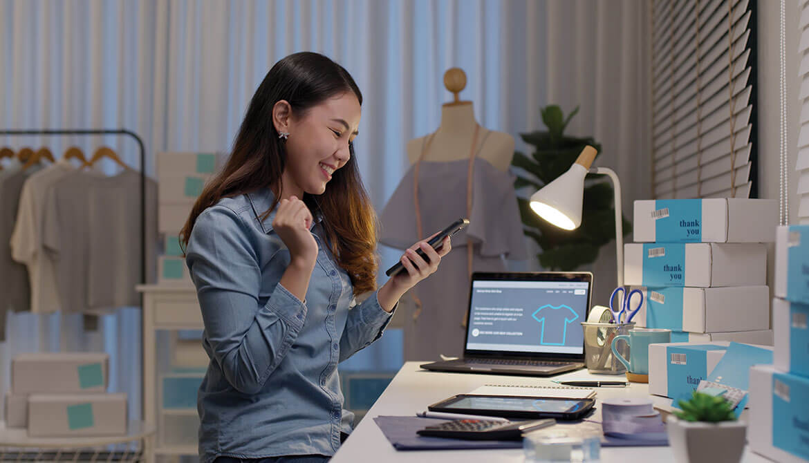 young female Asian small business owner makes a mobile deposit on smartphone while working at desk shipping packages