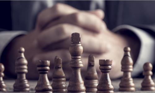 businessman with hands folded behind group of wooden chess pieces