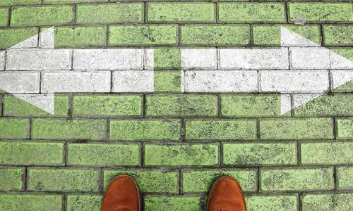 person in brown dress shoes standing on a green brick crossroad with white arrows pointing left and right