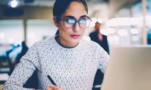 businesswoman wearing glasses writing in notebook while looking at laptop screen