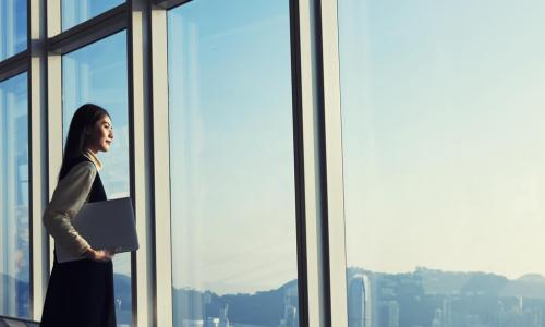 Young Asian millennial businesswoman holding laptop looks out high office windows