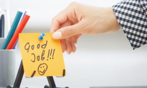 manicured hand reaching out to touch yellow good job sticky note on work desk