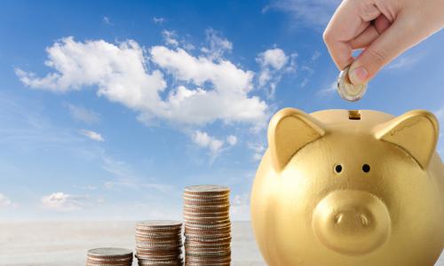 hand depositing coin in piggy bank next to stacks of coins on blue sky