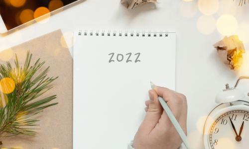 woman’s hand writing 2022 on notebook laying on desk surrounded by alarm clock phone and crumpled papers