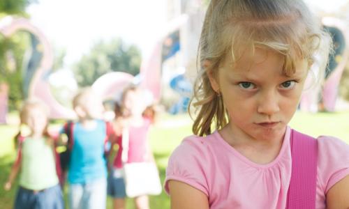 young girl stands by herself with arms folded defensively at playground park while group of 3 children exclude her