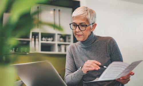 mature woman in glasses studies a paper printout while also looking at her laptop