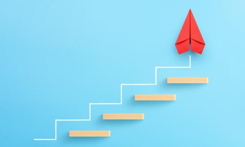 red paper airplane points upward as it progresses up wood block stairs