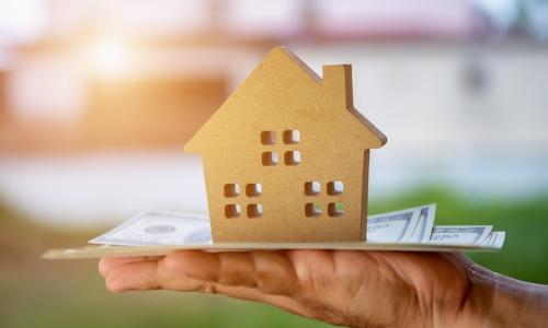 wooden cut-out of a house sitting on a stack of US dollars on an outstretched hand