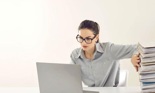 businesswoman working on a laptop while pushing a giant stack of folders and papers off the side of the table