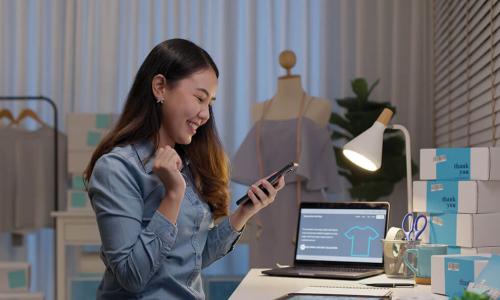 young female Asian small business owner makes a mobile deposit on smartphone while working at desk shipping packages