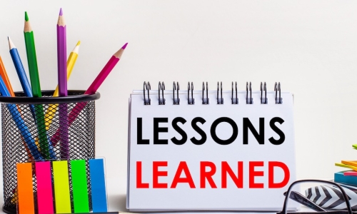 glasses and colored pencils with office supplies in front of notebook that reads LESSONS LEARNED