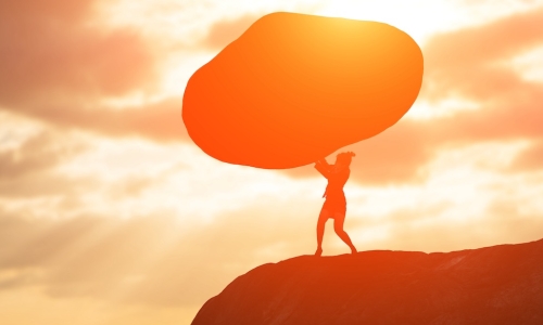 ambitious woman pushing up large boulder while standing on rocky outcrop