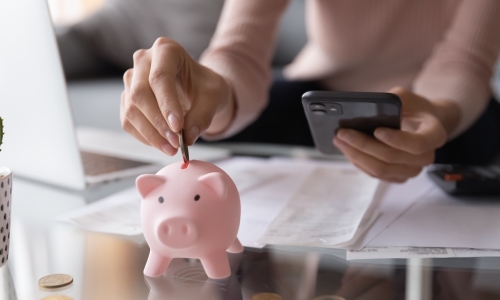 woman dropping a coin into a piggy bank while working on budget at her desk