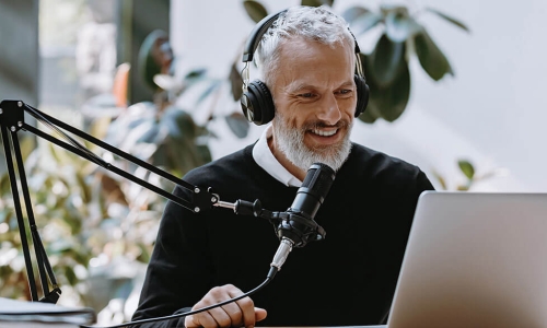 older male professional wears headphones and uses a professional microphone while recording on his laptop